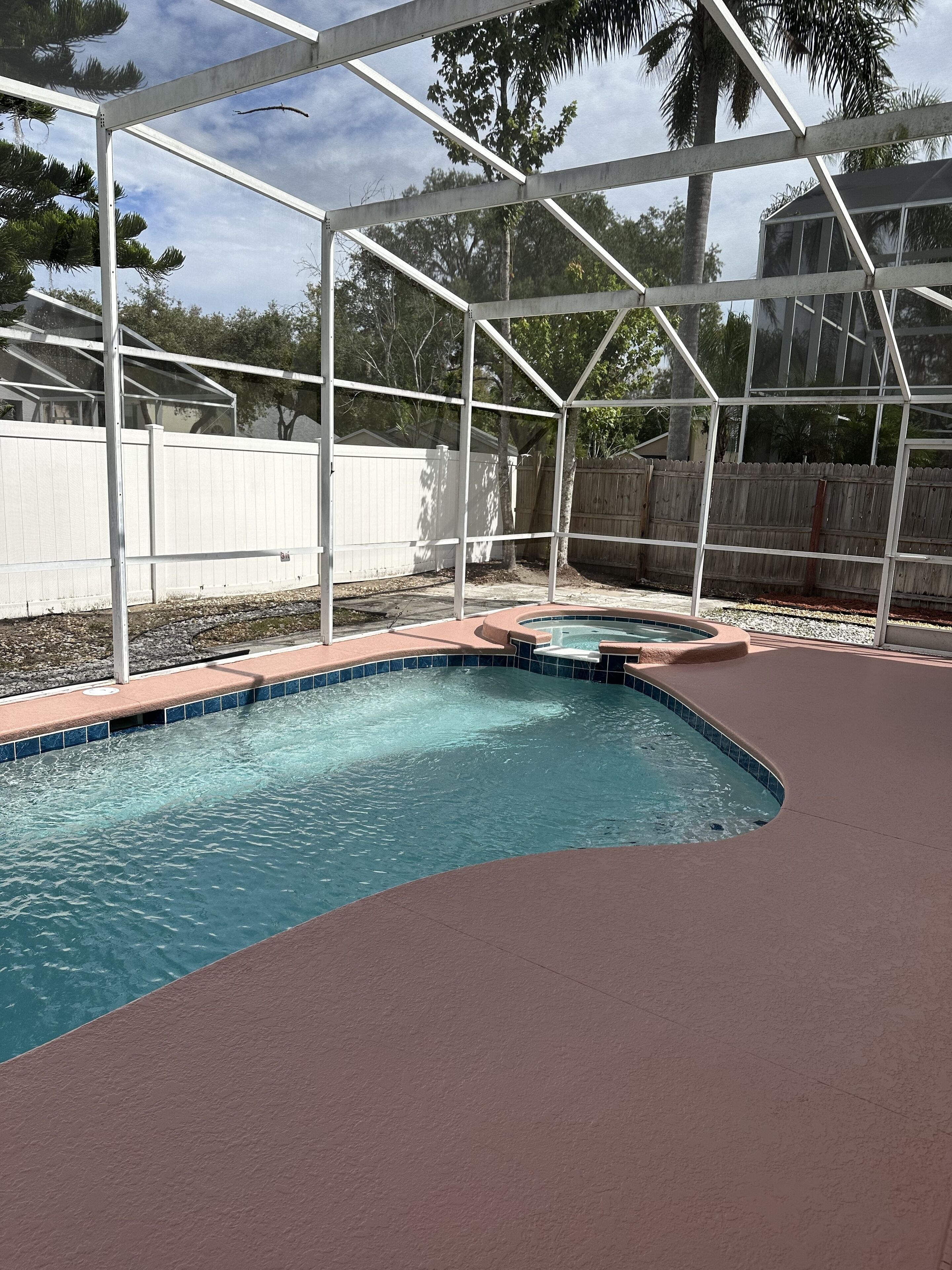 A swimming pool with a curved design, surrounded by a covered area with a metal frame and a white fence in the background.