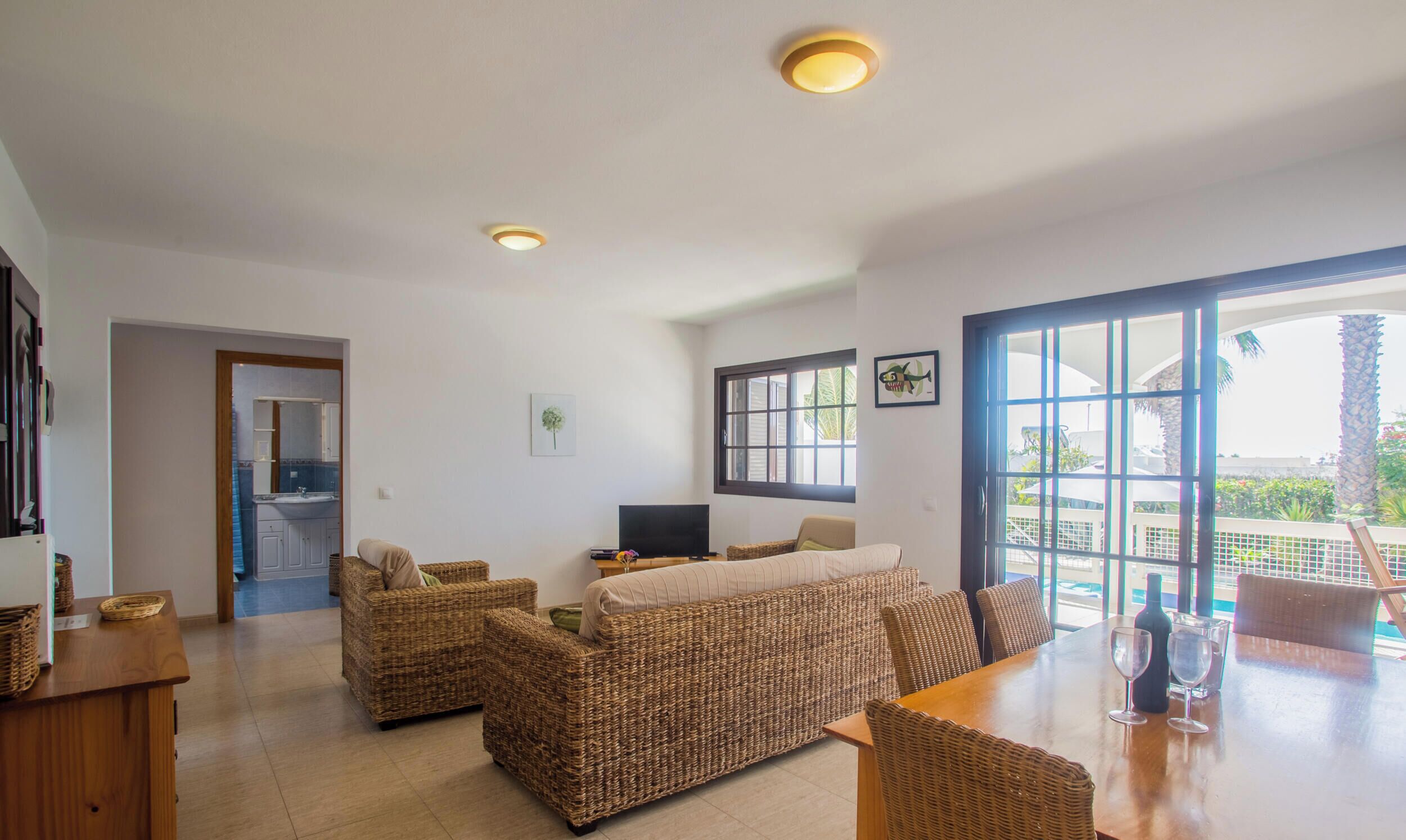 A spacious living area with wicker furniture, a dining table with glasses, and a large window offering a view of the outdoors.