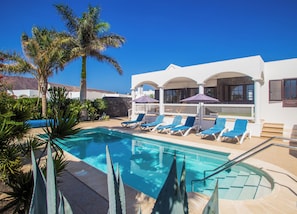 Una piscina con sillas de descanso, palmeras y un edificio blanco con ventanas arqueadas.
