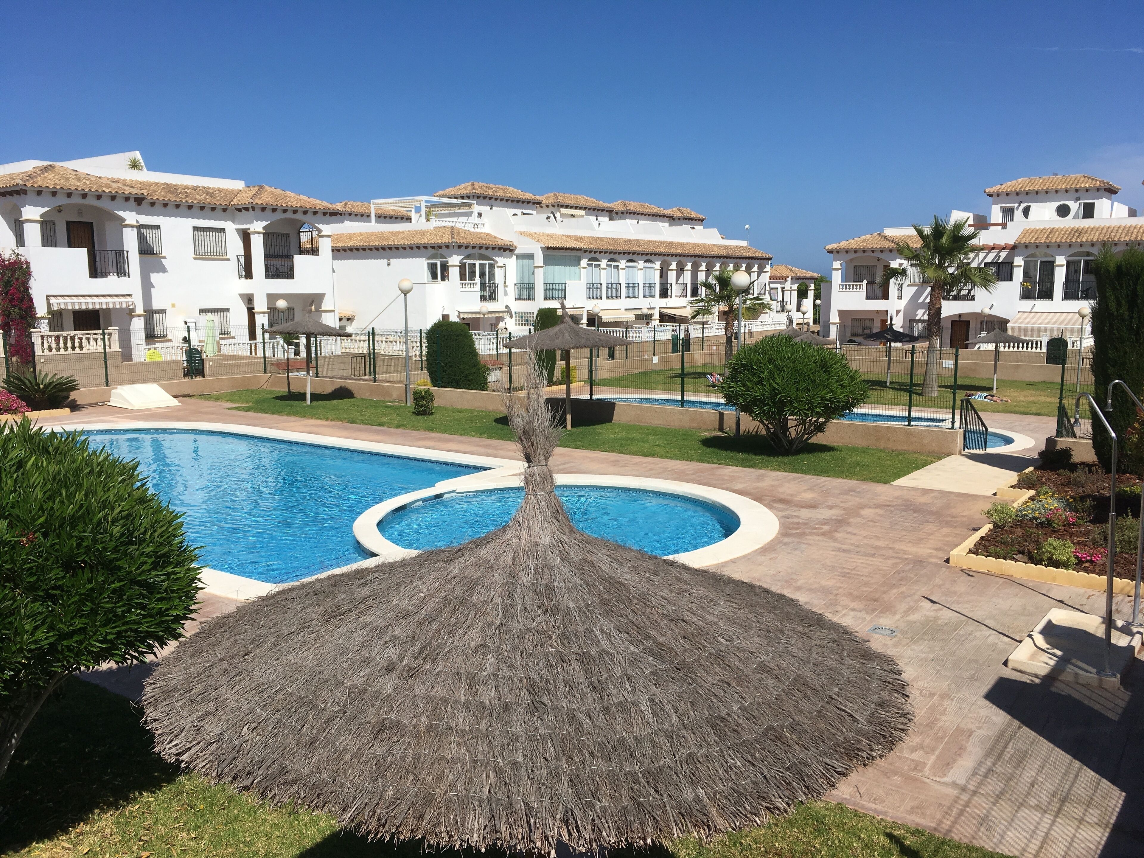 Una piscina con sombrilla de paja, rodeada de césped y plantas cuidadas, con edificios blancos de techos de tejas al fondo.