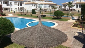 Una piscina con ombrellone di paglia, circondata da erba e piante curate, con sullo sfondo edifici bianchi con tetti in tegole.