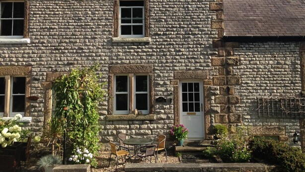 A stone building with a tiled roof, multiple windows, and a small outdoor seating area with chairs and a table.