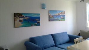 A room with a blue sofa, a small table, and two paintings of coastal scenes on the wall.