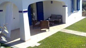 A covered outdoor seating area with a sofa and chairs, a tiled floor, and a grassy lawn.