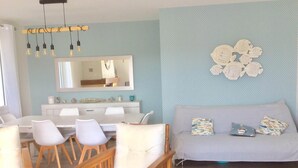 Un salon moderne avec un coin repas, caractérisé par un mur bleu clair, un grand miroir, une table à manger en bois avec des chaises blanches et un canapé gris agrémenté de coussins à motifs.