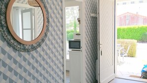Un couloir avec un papier peint à motifs, un miroir rond, un luminaire au plafond et une porte ouverte donnant sur une pièce où se trouve un four à micro-ondes.