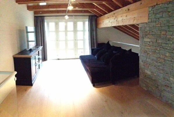 Una habitación con techo de madera, pared de piedra, sofá oscuro con cojines, un televisor sobre un mueble y un ventanal con cortinas.