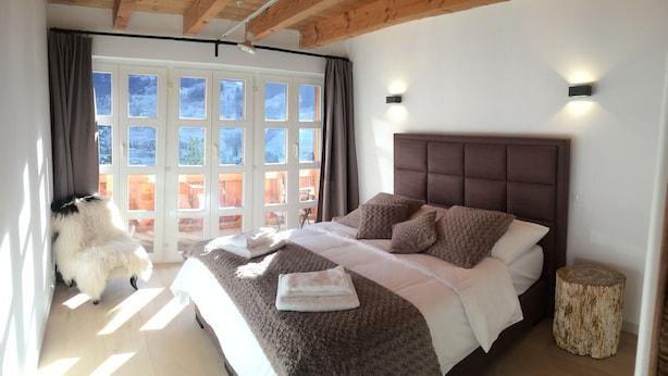 Ein Schlafzimmer mit einem großen Bett, einem hölzernen Kopfteil, einem kleinen runden Holzhocker und Blick auf eine Holzterrasse durch ein Fenster.