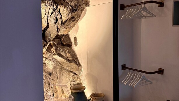 A room with a stone wall archway, a bed, a hanging clothes rack, and a small table with pots.