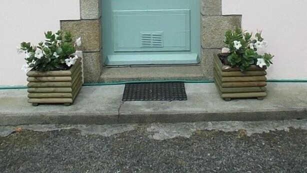 Una porta color petrolio con stipite in pietra, una piccola luce sopra e due vasi con fiori bianchi ai lati.