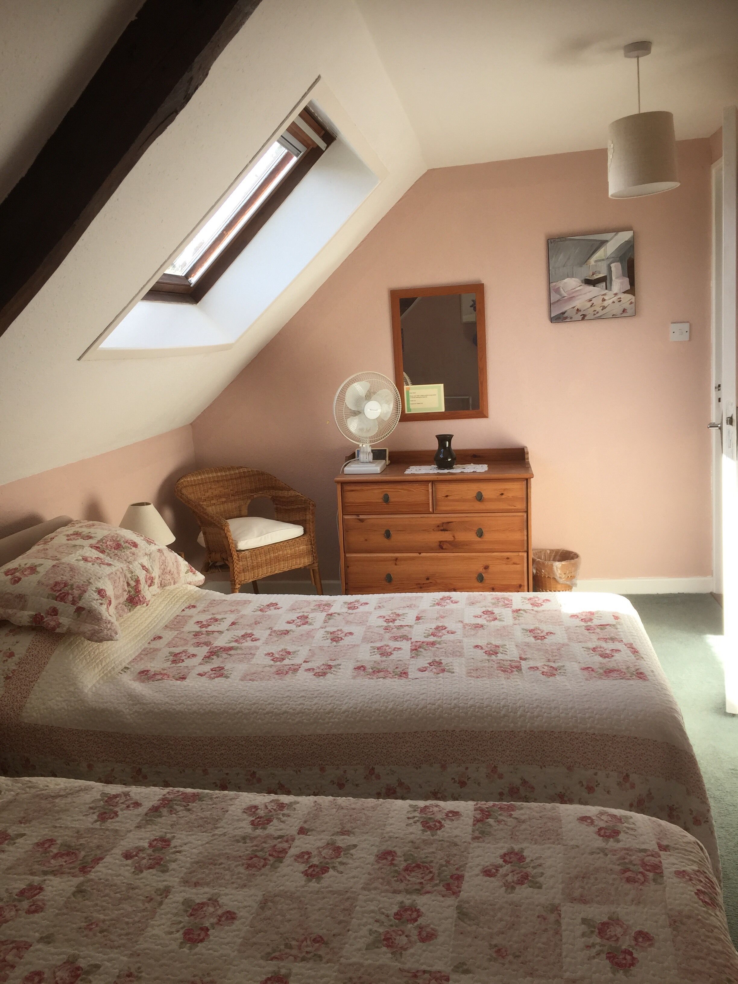 A bedroom with a sloped ceiling, a skylight, a bed with floral bedding, a wooden dresser, a chair, and a painting on the wall.