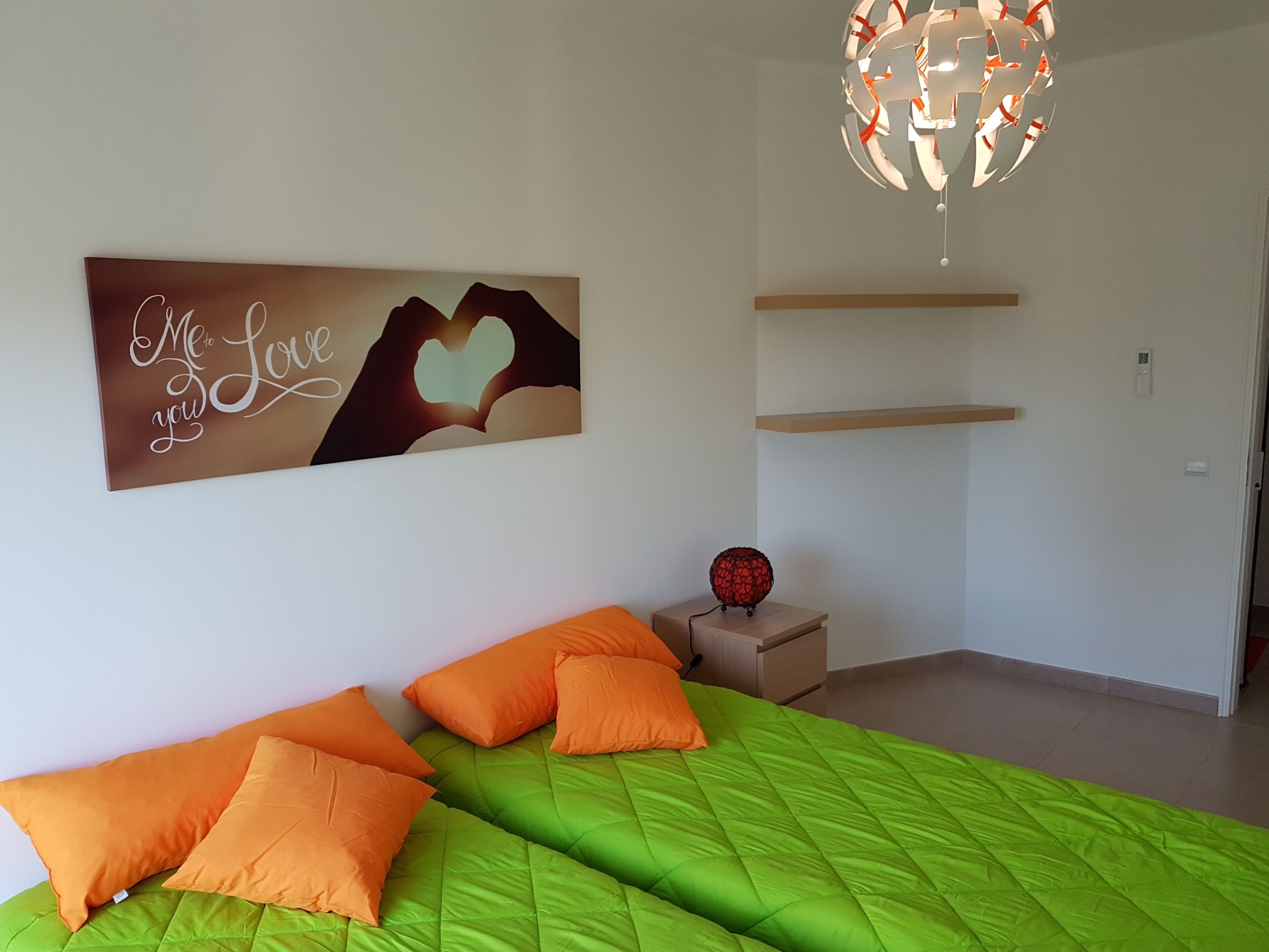Een slaapkamer met twee bedden, oranje kussens, een groene gewatteerde plaid en een wanddecoratie met een hartmotief, plus een hanglamp.