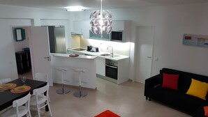A modern kitchen with white cabinets, a central island with stools, a microwave, and a stove. There is a dining area with a table and chairs, and a black sofa with red and yellow cushions. - Fuzeta beach apartment 1 - Free Wifi (Olhao)