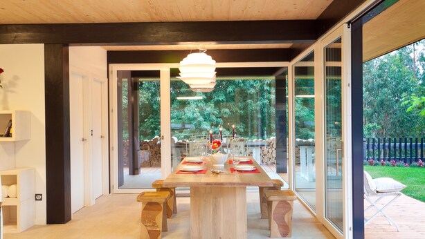 A modern dining area with a wooden table set for four, large glass doors leading to a garden, and a hanging light fixture. - House with pool 8 minutes from the beach, free wifi (Ea)