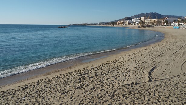 Ein sandiger Strand mit Fußabdrücken, ein ruhiges Meer und Gebäude im Hintergrund.