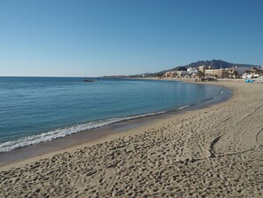 Une plage de sable avec des empreintes, une mer calme et des bâtiments en arrière-plan.