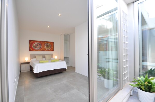 A modern bedroom with a bed, bedside lamps, a large abstract painting, and a view through a sliding glass door.