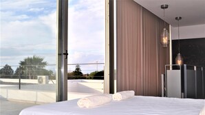 Une chambre d’hôtel moderne avec un grand lit, deux serviettes roulées, une porte vitrée donnant sur un balcon et une vue sur l’extérieur.