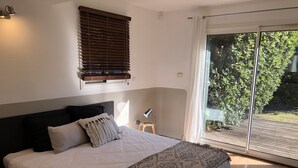Un dormitorio con una cama grande, una mesita de noche, una lámpara, una ventana con persianas y vistas al exterior.