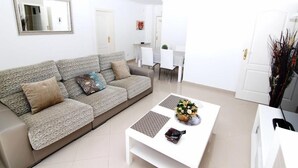 A modern living room with a grey sofa, a white coffee table with a flower vase, and a dining area in the background.