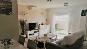 A modern living room with a flat-screen TV, white furniture, a dining area with a table and chairs, and a view of a green outdoor space.