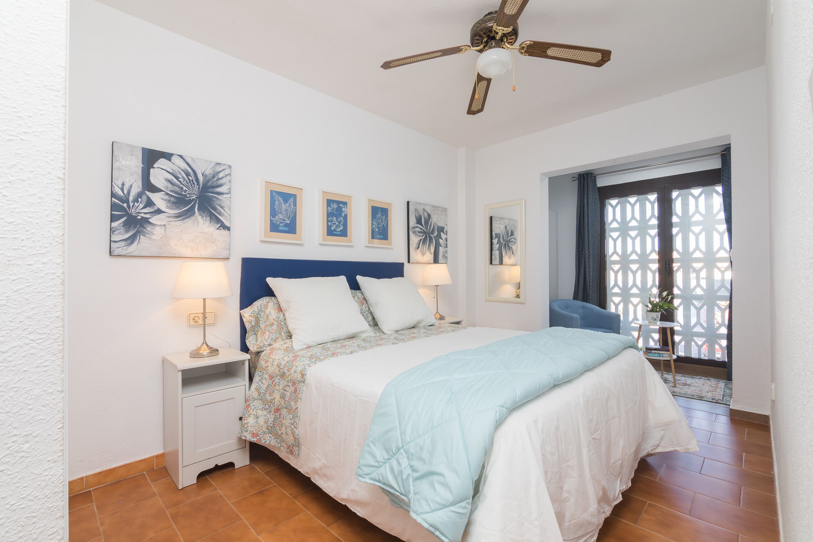 A bedroom with a bed, nightstands, wall art, a ceiling fan, and a view through a window with a lattice screen.
