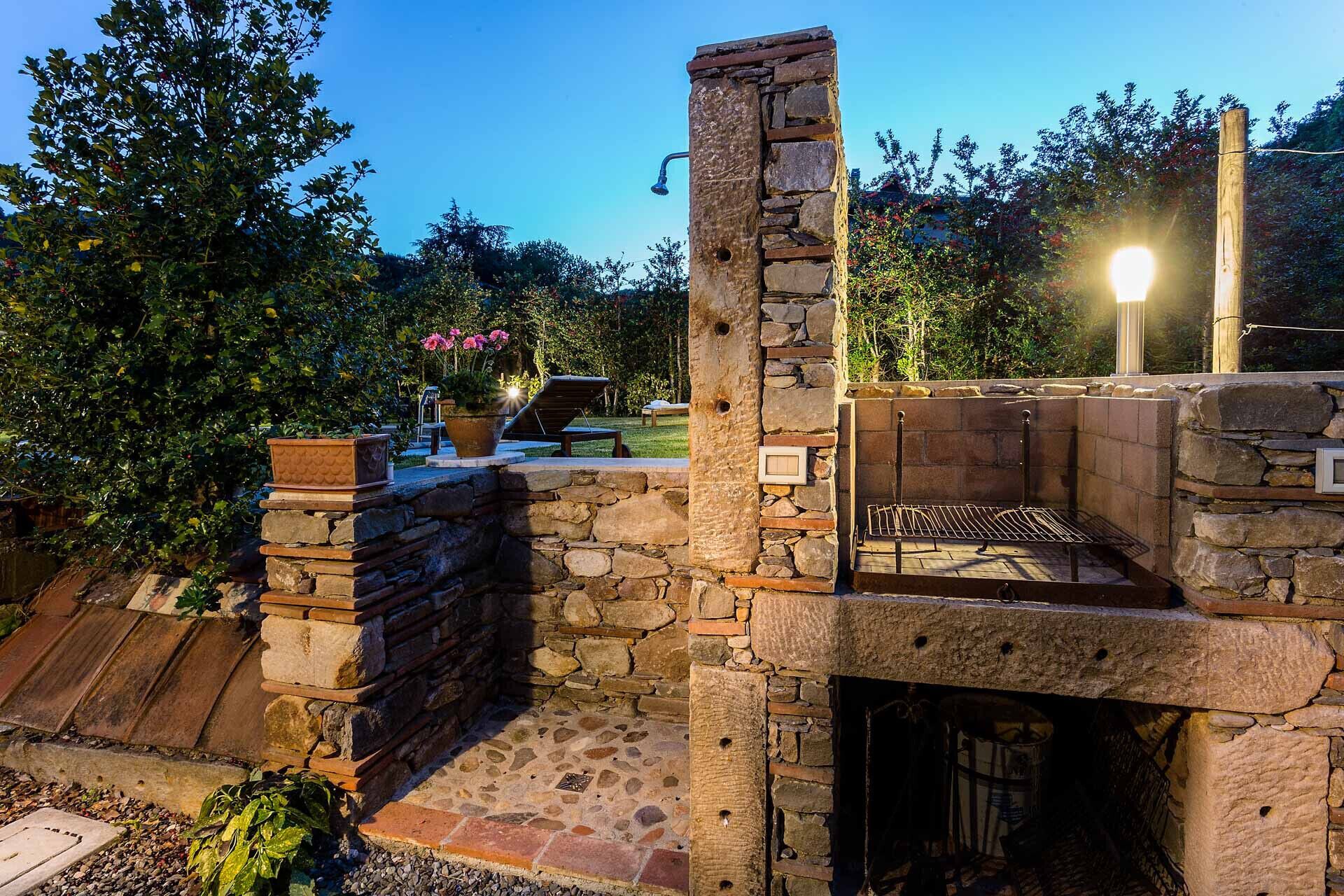 A stone-built outdoor kitchen with a grill, surrounded by greenery and a wooden deck.