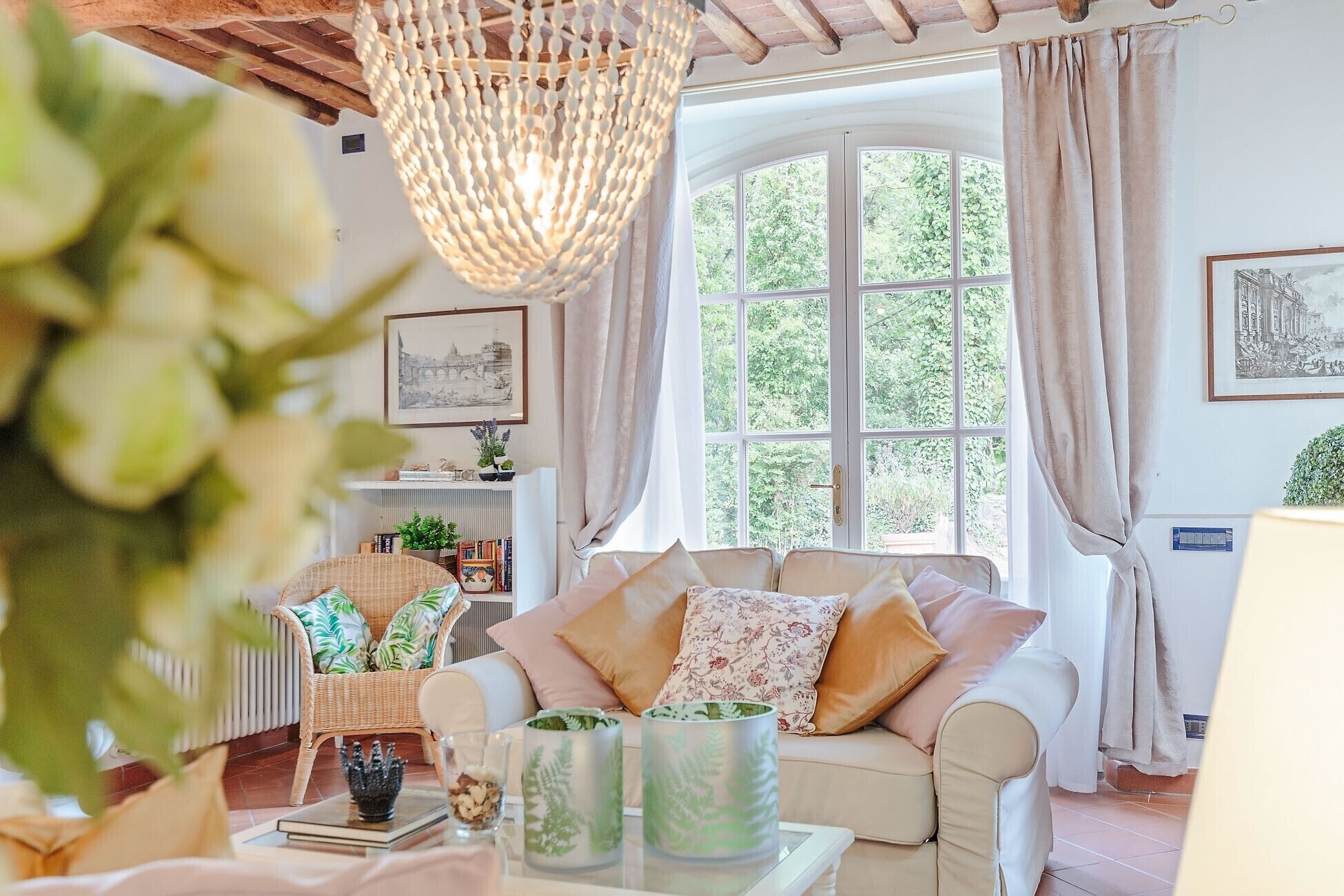 A cozy living room with a large window, a wicker chair, a sofa with floral cushions, and decorative vases on a coffee table.