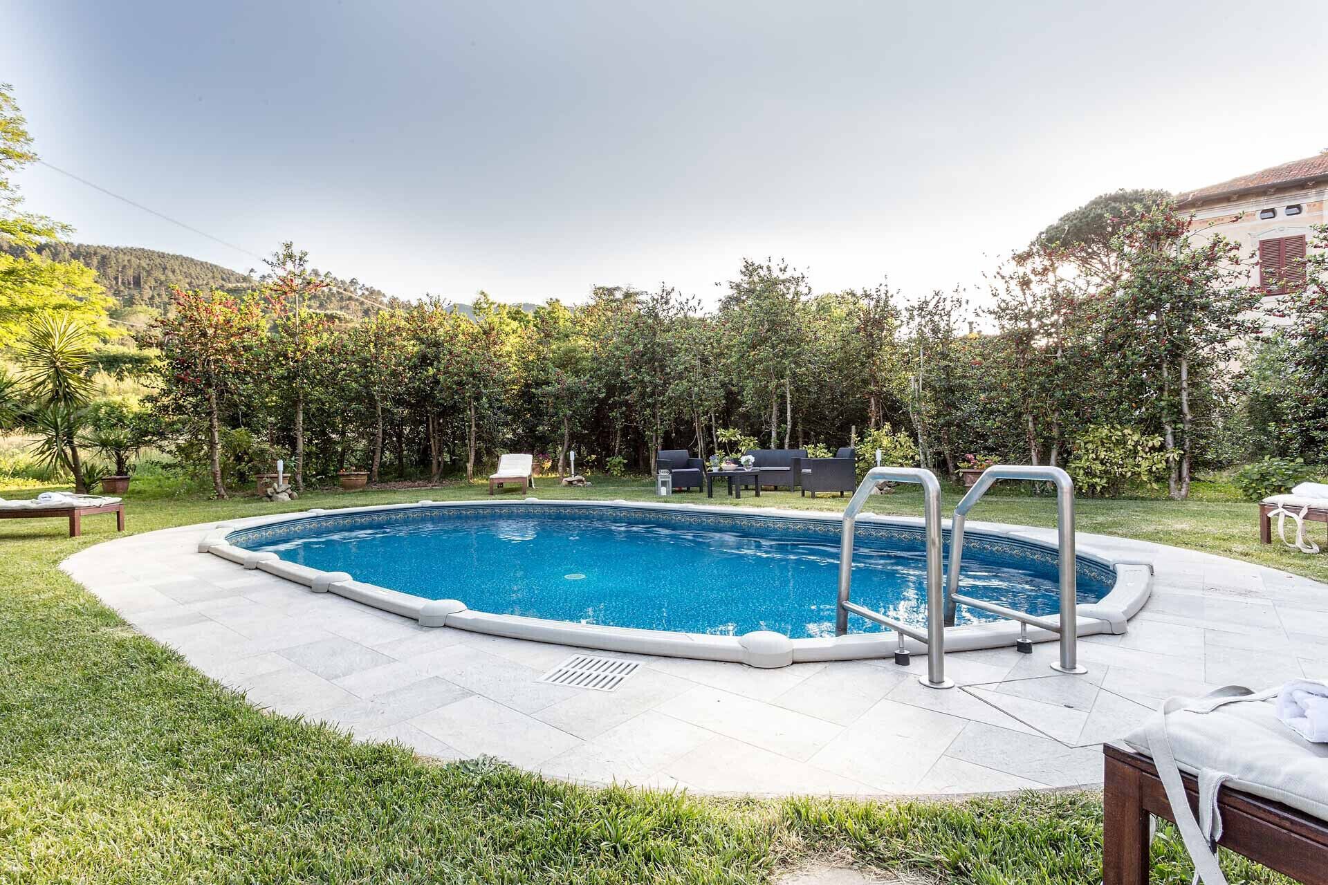 A rectangular swimming pool with a metal ladder, surrounded by a grassy area with a few lounge chairs and a small table.