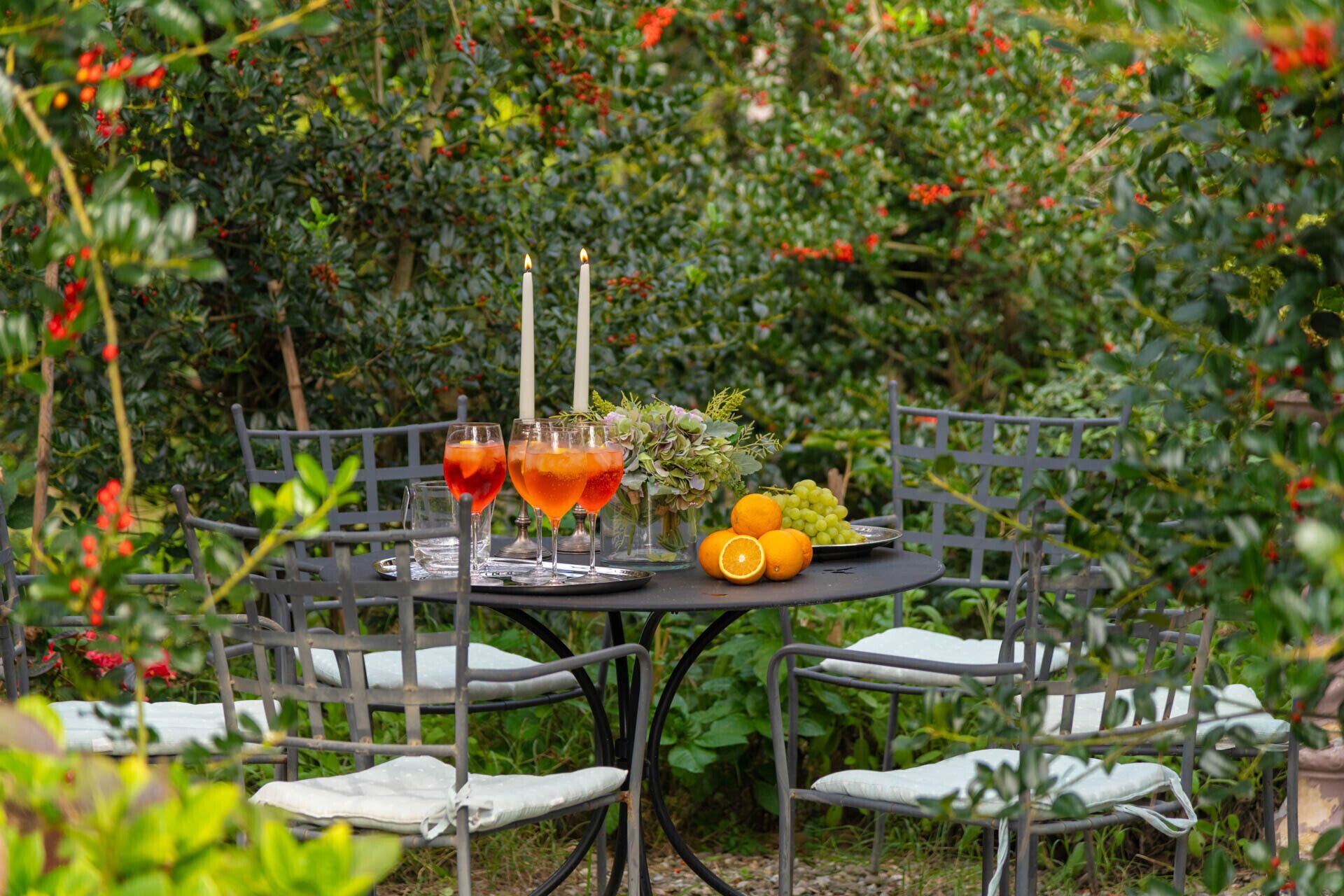 Una mesa de jardín con estructura metálica, cojines blancos y una bandeja con bebidas, fruta y un jarrón con follaje.