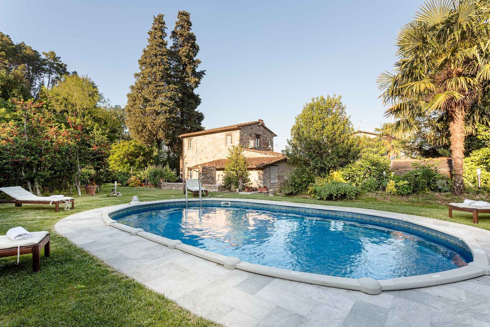 A swimming pool with lounge chairs, surrounded by a stone house and lush greenery.
