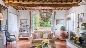 A cozy living room with a sofa, a coffee table with fruits and tea set, a bookshelf, and a large window with a view of greenery. - A secret sweet idyllic retreat for 2 couples with private pool & air conditionin (Pieve di Compito)