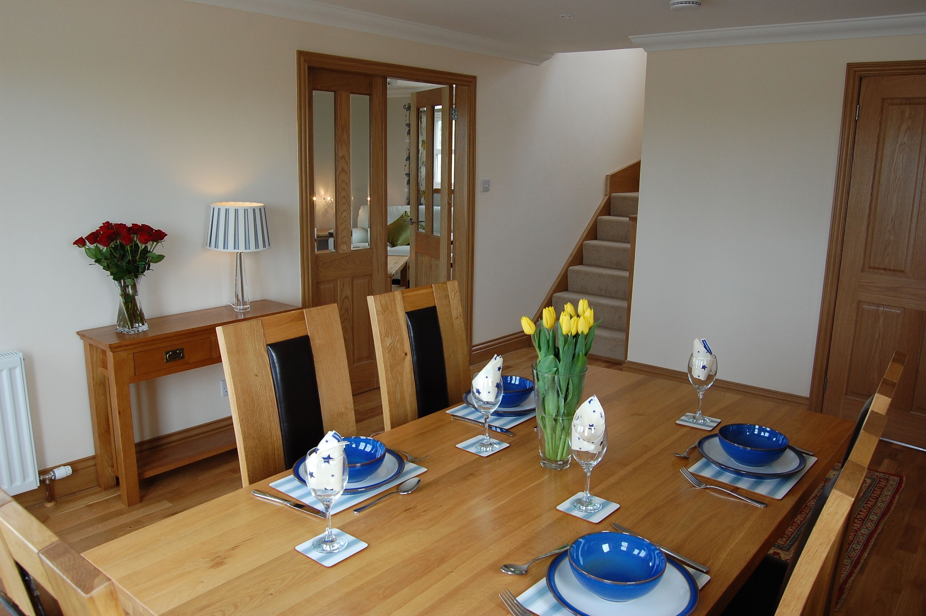 A dining area with a wooden table set for six, blue plates, and a vase of yellow tulips.