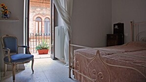 Un dormitori amb un llit, una cadira, una planta en test i una vista d'un edifici a través d'una porta oberta.