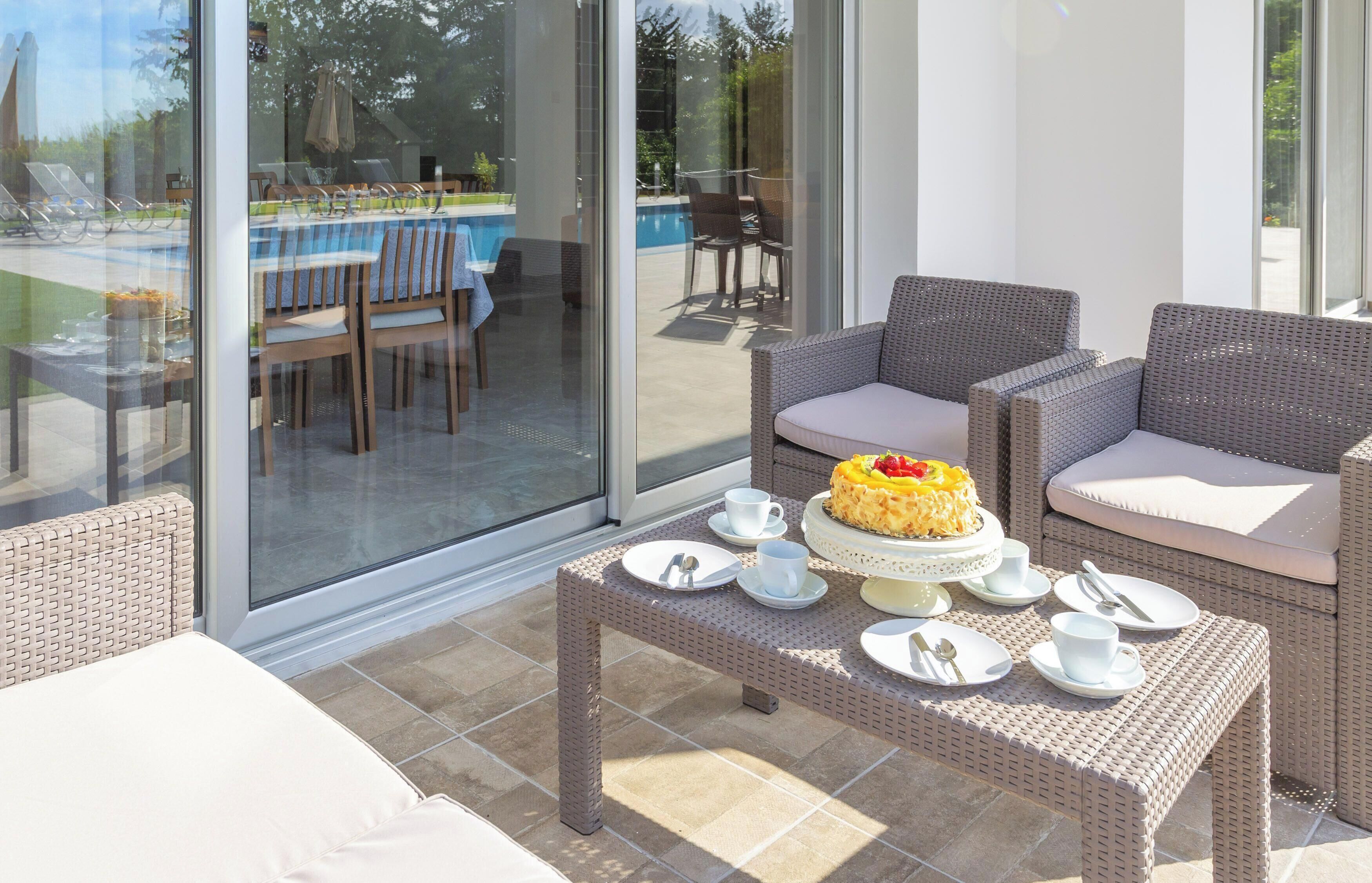 Une terrasse ensoleillée, meublée de mobilier en osier, avec une table à manger dressée pour deux personnes, et une vue sur une piscine extérieure et un espace repas.