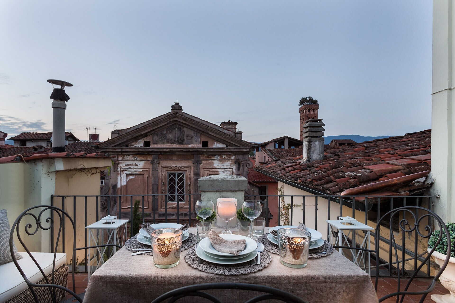 A table set for a meal with candles, plates, and glasses, overlooking a historic building with a tiled roof.