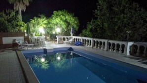 Zona de piscina nocturna con suelo de baldosas, sillas de descanso y barandilla decorativa.