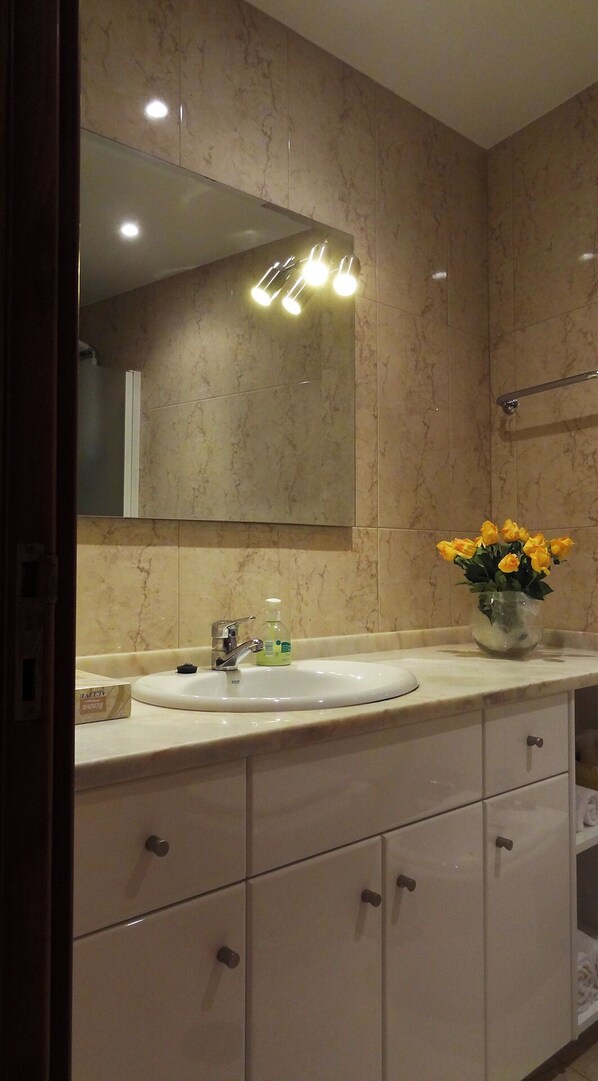 Une salle de bain avec un meuble-lavabo en marbre, un évier, un miroir et un vase rempli de fleurs jaunes.