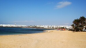 A beach with a clear sky, a sandy shore, and a white building complex in the background. - Beautiful beachside apartment with stunning sea views. Seven pools on complex! (Costa Teguise)