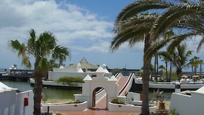 A resort entrance with a white archway, palm trees, and a view of the sea. - Beautiful beachside apartment with stunning sea views. Seven pools on complex! (Costa Teguise)