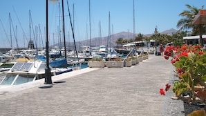 A marina with boats docked, a paved walkway, and a mountain in the background. - Beautiful beachside apartment with stunning sea views. Seven pools on complex! (Costa Teguise)