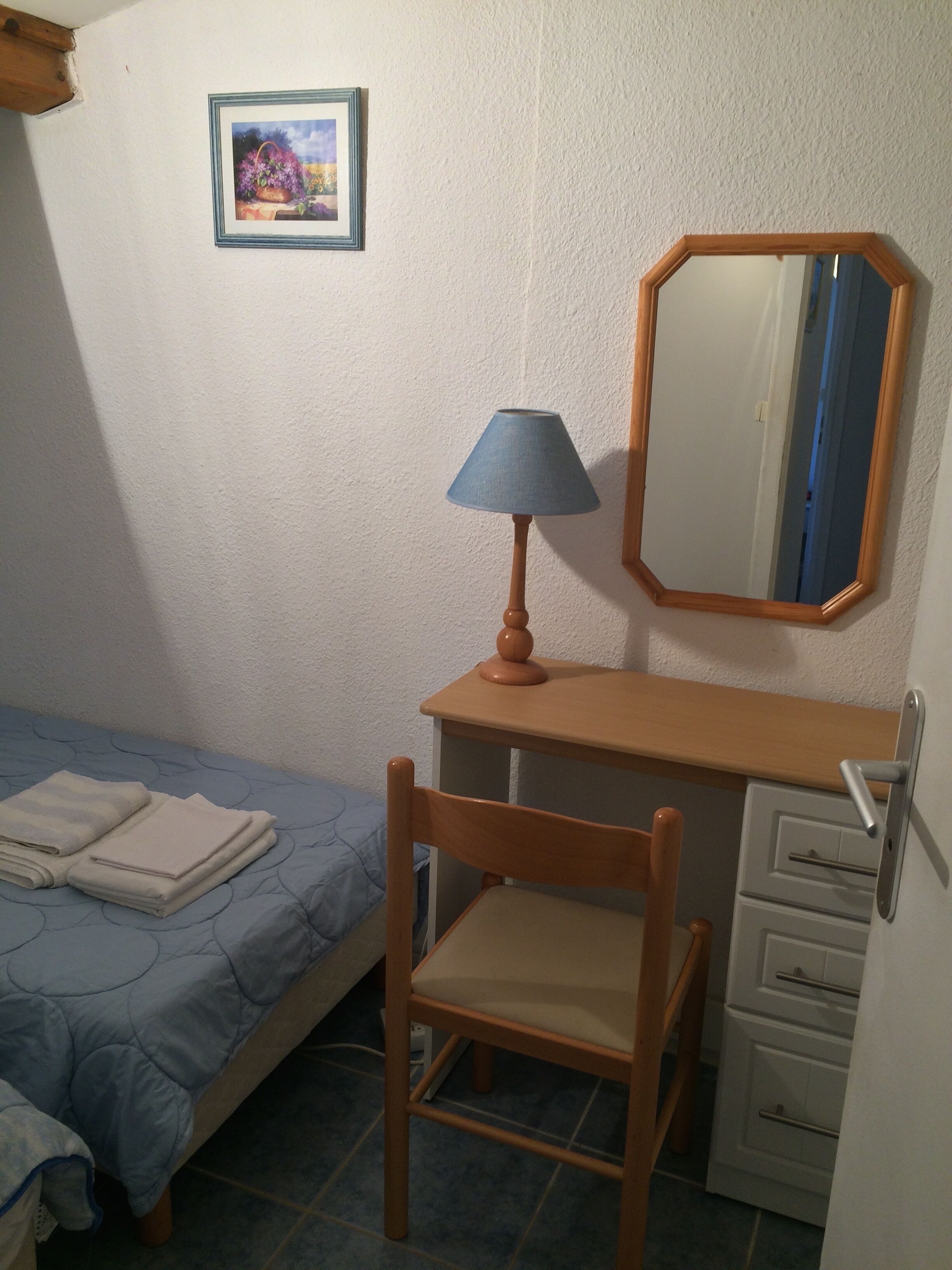 Ett litet sovrum med en enkelsäng, ett skrivbord med en stol, en lampa, en spegel och en byrå.