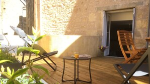 Um pátio externo com piso de madeira, duas poltronas de descanso, uma mesa pequena com um copo de suco de laranja e uma porta que dá para um espaço interno.