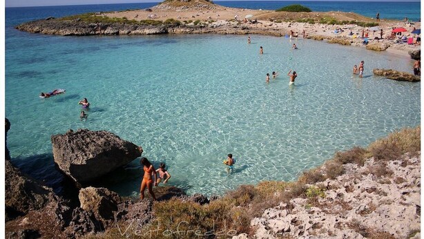 Een kuststrand met helder turquoise water, rotsachtig terrein en een kleine heuvel op de achtergrond.