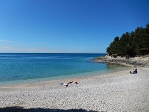 A pebbled beach with clear blue water, a rocky shoreline, and a person sitting on the sand. - Deluxe apartment 8 + 2, with private pool, 100m from the beach (Pula)
