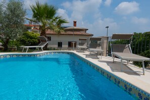 A swimming pool with lounge chairs, a palm tree, and a building with a tiled roof in the background.