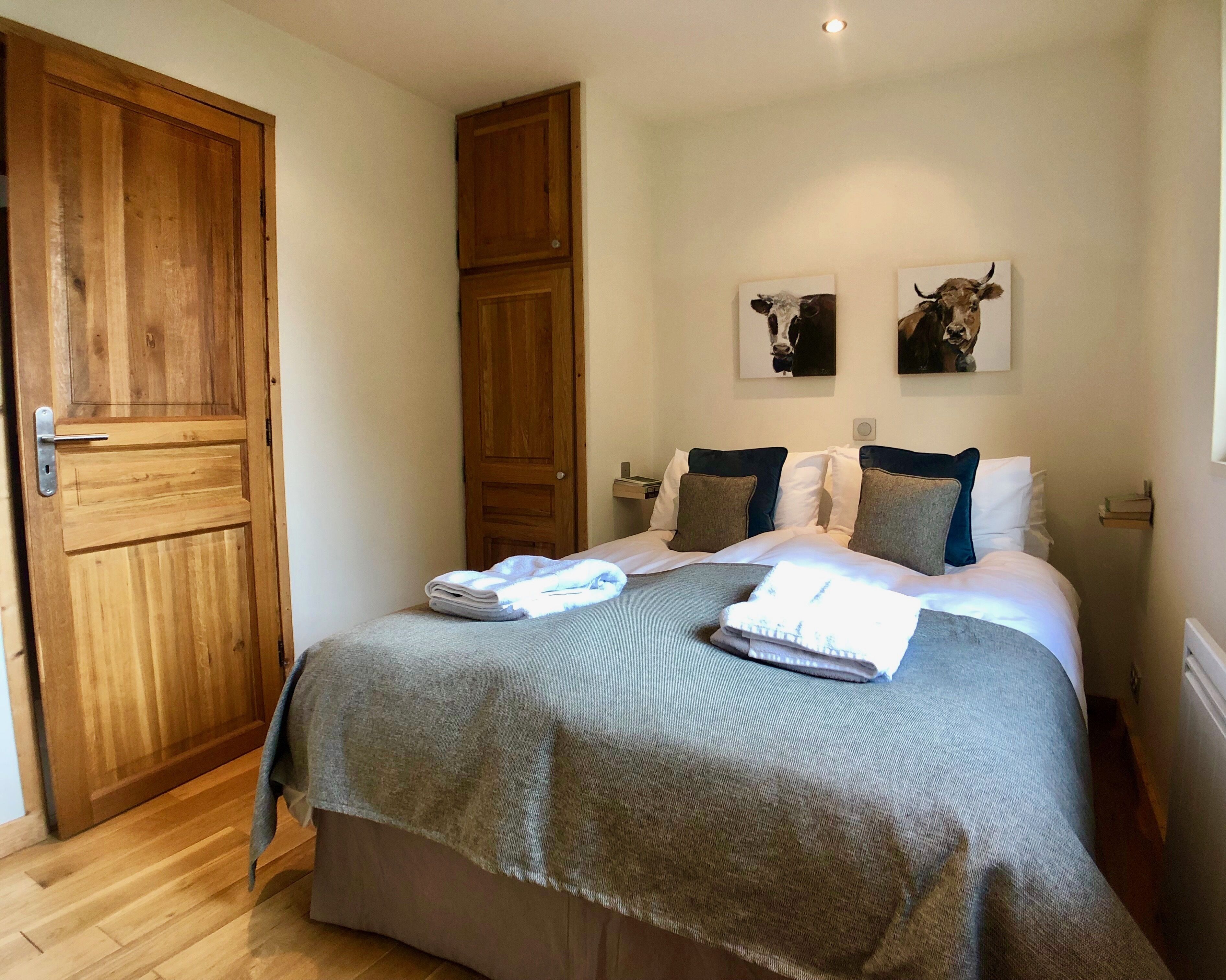 A neatly made bed with a grey blanket and two pillows, a wooden wardrobe, and two cow-themed wall art pieces.