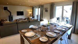 Zona pranzo con tavolo di legno apparecchiato per sei persone, un divano e una TV sullo sfondo.