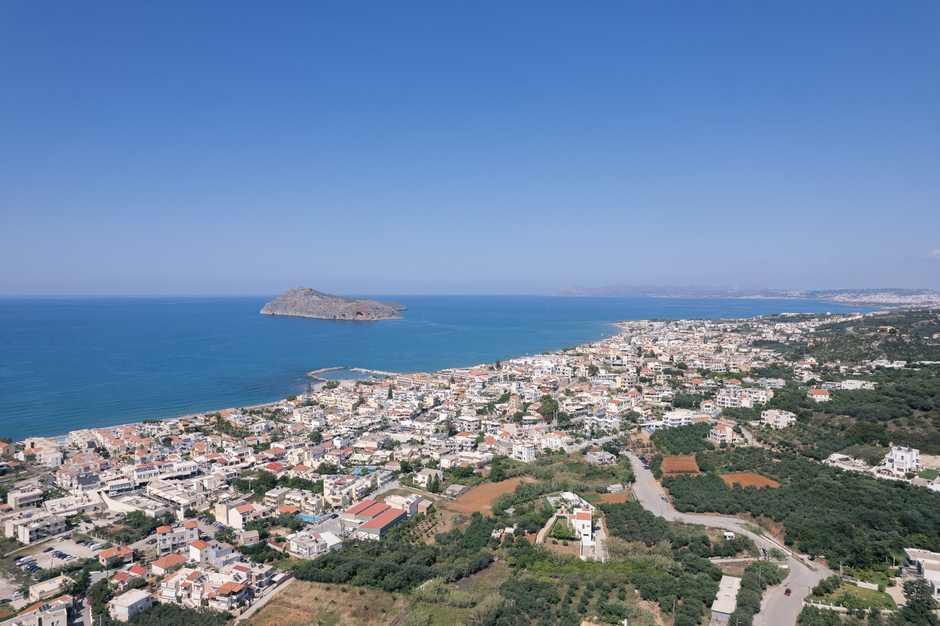 Uma cidade costeira com mistura de edifícios residenciais e áreas abertas, uma ilha rochosa proeminente no mar, e um céu azul límpido.