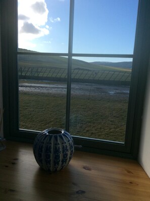 A room with a large window overlooking a field and distant hills. A ceramic vase is placed on a wooden table in the foreground.